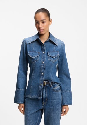 Femme portant une chemise en jean bleue boutonnée avec de larges poignets et un jean bleu assorti sur un fond blanc uni.