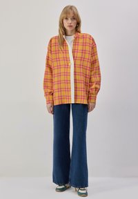 Veste surdimensionnée à carreaux jaune et rose avec une fermeture à boutons blancs, associée à un jean bleu foncé évasé et des baskets avec des accents verts.