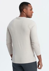 Man som står på sidan med en enkel, ljusbeige långärmad skjorta och mörka byxor mot en vit bakgrund.