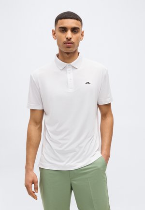 Mann trägt weißes kurzärmeliges Poloshirt mit schwarzem Logo und hellgrünen Hosen, steht mit einer Hand in der Tasche vor einem einfarbigen Hintergrund.