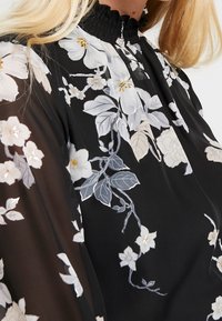 Svart blus med blommig stickning i vitt och ljusblått, med en smockad halsringning och genomskinliga, flödande ärmar.