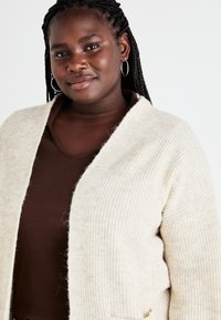 Crème kleurige cardigan met een zachte, textuur gebreide stof. Open voorkant ontwerp met een relaxte pasvorm, gedragen over een bruine rib gebreide top.