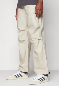 Pantalones cargo beige de corte holgado, con múltiples bolsillos, textura visible y aberturas en los tobillos ajustables, combinados con zapatillas blancas.