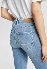 Jeans de tiro alto en azul claro, con un parche de cuero, bolsillos traseros y detalles de costura visibles. La tela es de mezclilla con un acabado ligeramente desgastado.