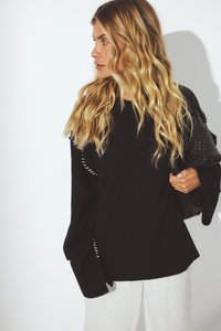 Schwarzer gestrickter Pullover mit weiten Ärmeln, geripptem Saum und seitlichen Schlitzen, kombiniert mit leichten, strukturierten Hosen. Stylisch ergänzt mit einem gemusterten Accessoire.