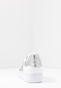Bílé tenisky Adidas s metalicky stříbrným patním dílem a logem, zobrazené ze zadní strany na bílé ploše a pozadí.