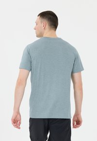 Mies, jolla on lyhyet hiukset ja korvakoru, pukeutuneena vaaleansiniseen t-paitaan ja mustiin shortseihin, nähtynä takaa yksinkertaista valkoista taustaa vasten.