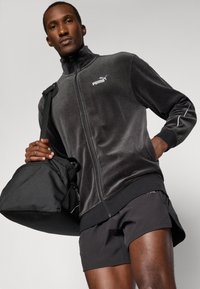 Μαύρο βελούδινο μπουφάν με φερμουάρ και ψηλό γιακά, με λευκό λογότυπο Puma. Συνδυάζεται με μαύρες βερμούδες και κρατάει μια μαύρη τσάντα duffel.