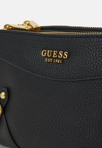 Černá kabelka z texturované kůže se zlatě tónovanými kovovými prvky a zipy. Na přední straně se nachází logo s nápisem "GUESS EST 1981".