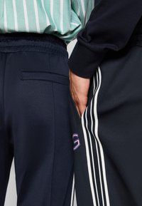 Jogging en bleu marine avec taille élastique, détails de poches et accents de rayures blanches sur le côté. Haut rayé vert partiellement visible.