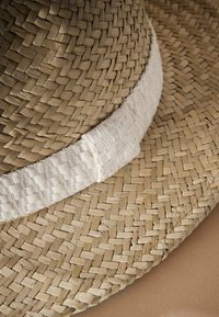 Chapeau de paille à large bord avec une texture étroitement tissée de couleur naturelle beige clair, souligné par un ruban en tissu doux de couleur claire. Bords lisses visibles.
