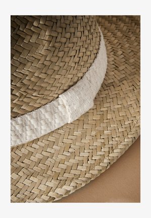 Vid brimmed stråhat med en tæt vævet tekstur i naturlig tan, fremhævet af et blødt, lyst farvet stofbånd. Glatte kanter er synlige.