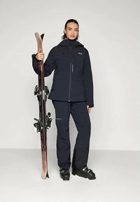 Veste de ski bleu marine foncé et pantalon assorti, accompagnés de chaussures de ski noires. Le mannequin tient un ski avec des fixations noires et une finition imitant le bois.
