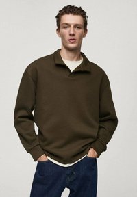 Pullover marrone, comodo, con colletto, realizzato in materiale morbido. Indossato sopra una camicia bianca e abbinato a jeans di denim blu.