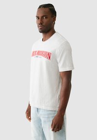 Šviesiai pilkas trumpomis rankovėmis T-shirts, pagamintas iš medvilnės, su išryškintu raudonu išsiuvinėtu logotipu krūtinėje ir laisvu siluetu.