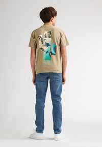 Beige bomulds-t-shirt med stort grafisk design på ryggen, der indeholder blå og jordfarvede toner, med korte ærmer og rund halsudskæring.