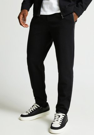 Homme portant un pantalon de jogging noir, des baskets noires et blanches, une chemise blanche et une veste zippée noire, les mains dans les poches.