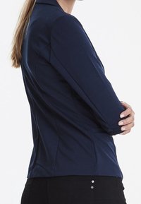 Mörkblå skräddarsydd blazer med långa ärmar, slätt tyg, strukturerade axlar och subtila sömnadsdetaljer, bärs över svarta byxor.