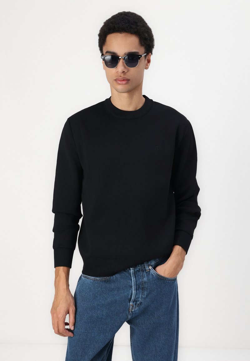 Felpa nera in pile con girocollo, dotata di polsini e orlo a costine, abbinata a jeans in denim blu. Il modello indossa occhiali da sole scuri.