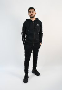 Chándal deportivo negro compuesto por una sudadera con cremallera y pantalones estrechos, que presenta detalles en blanco con logotipos y texto a lo largo de las mangas y las piernas.