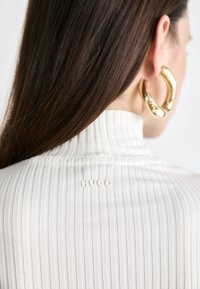 Donna con lunghi capelli castani che indossa un dolcevita bianco a coste con la scritta "HUGO" e grandi orecchini a cerchio dorati, di spalle su uno sfondo bianco.