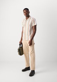 Chemise à rayures à manches courtes en crème avec des rayures multicolores, pantalon beige et bottines noires à la cheville. Portant un petit sac vert olive.