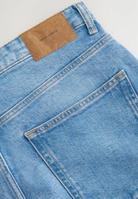 Jeans din denim de culoare albastră deschisă, cu o etichetă din piele maro pe talie, pe care este gravat "DENIM SUPPLY". Țesătura are o textură moale.