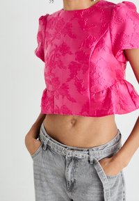 Haut court en satin rose avec motif floral texturé, manches courtes bouffantes et ourlet à volant, porté avec un jean gris taille haute.