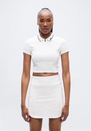 Tricou polo alb, scurtat, cu fermoar la gât și detalii negre la guler, asortat cu o fustă mini albă, mulată, cu textură fină.