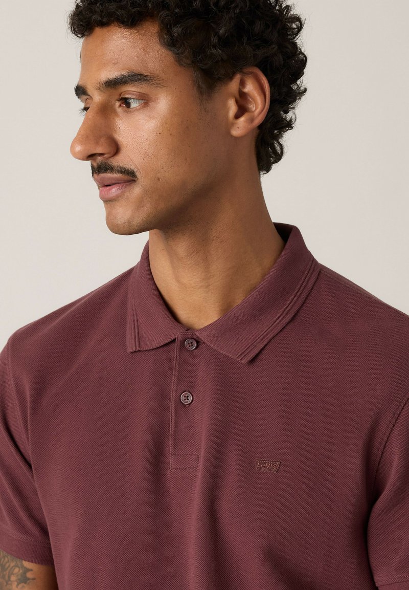 Hombre con cabello rizado y bigote, vistiendo una camiseta tipo polo granate de Levi's, mirando hacia su izquierda sobre un fondo liso.
