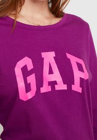 Lila sweatshirt med djärva rosa bokstäver som stavar "GAP." Tyget verkar slät med en rund halsringning och avslappnad passform.