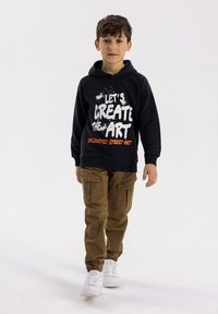 Svart hoodie med vit och orange grafisk text, kombinerad med beige cargobyxor och vita sneakers. Utanför har en avslappnad stil.