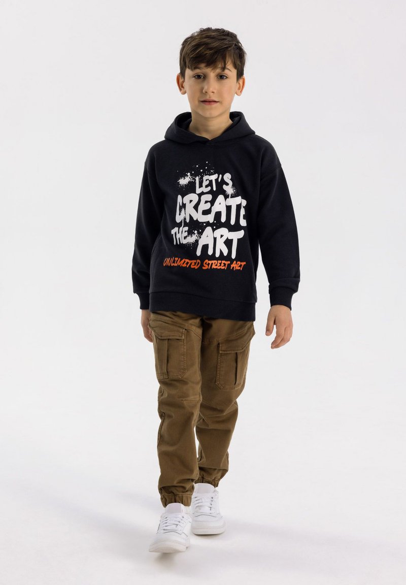 Svart hoodie med vit och orange grafisk text, kombinerad med beige cargobyxor och vita sneakers. Utanför har en avslappnad stil.