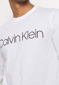 White long-sleeve T-shirt made of cotton. Features a large black "Calvin Klein" logo across the chest, crew neck design, smooth texture.