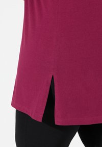Top magenta a maniche corte con orlo arrotondato e fessure laterali, realizzato in tessuto morbido. Indossato sopra leggings neri per un contrasto.