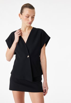 Donna con i capelli lisci tirati indietro che indossa una maglia nera a maniche corte con un solo bottone e una gonna nera abbinata, che guarda in basso di lato.