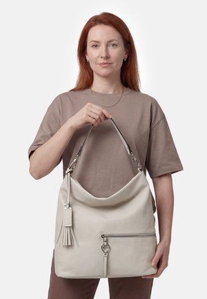 Mujer con cabello rojo que lleva una camisa topo, sosteniendo un bolso grande beige con cremallera y detalle de borla.