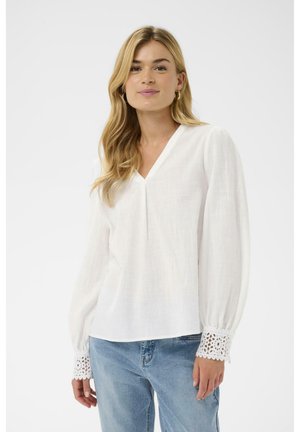 Biela blúzka s dlhým rukávom z ľahkej tkaniny, so zapínaním do V a čipkovaným lemom na manžetách. Kombinovaná s modrými džínsami.