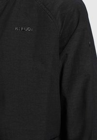 Veste noire avec une finition texturée, arborant un logo discret "Khujo" sur l'épaule. Le design présente une silhouette épurée et minimaliste.