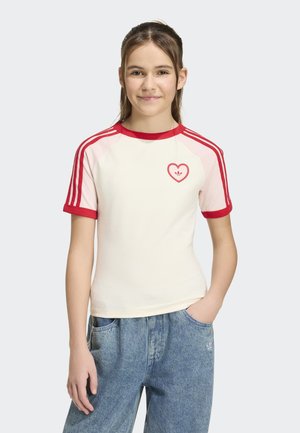 Fille portant un t-shirt blanc et rouge à manches courtes avec un logo en forme de cœur et un jean bleu en denim, souriante avec une main dans la poche.