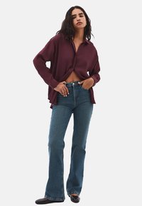 Camicia bordeaux a maniche lunghe con una vestibilità morbida, abbinata a jeans blu a campana aderenti. Tessuto liscio con colletto classico e una leggera lucentezza.