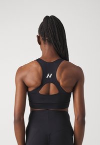Crni sportski grudnjak s dizajnom na racerback, s izrezanim detaljem na leđima i reflektirajućim logotipom. Glatki, rastezljivi materijal.
