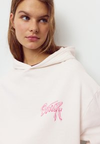 Felpa con cappuccio rosa chiaro in tessuto morbido, con un logo ricamato rosa decorativo sulla parte anteriore, dal taglio ampio e con cappuccio grande.