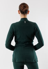 Maglietta a maniche lunghe verde scuro con collo alto, texture morbida, design aderente e una cucitura centrale, con un piccolo etichetta del marchio al collo.