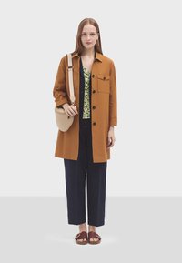 Dlhý hnedý kabát cez zelený kvetinový top, doplnený námorníckymi rebrovanými nohavicami. Ozdobený hnedými sandálmi a okrúhlou béžovou crossbody taškou.