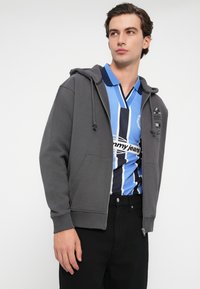 Szara bluza z kapturem na zamek błyskawiczny, z kieszeniami z przodu i ściągaczami przy mankietach, nałożona na niebieską i czarną polo w paski, połączona z czarnymi spodniami.