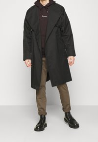 Czarny płaszcz trencz o luźnym kroju, z dwoma sznurkami, noszony na ciemnej bluzie z kapturem, w połączeniu z brązowymi spodniami i czarnymi butami na sznurowadła.