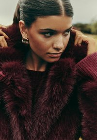 Burgundijas mākslīgās kažokādas mētelis ar teksturētu apdari, kuram ir masīvs adīts apkakle un zeltītas spirāļu auskari. Tuva plāna cilvēka kakla un sejas daļa.