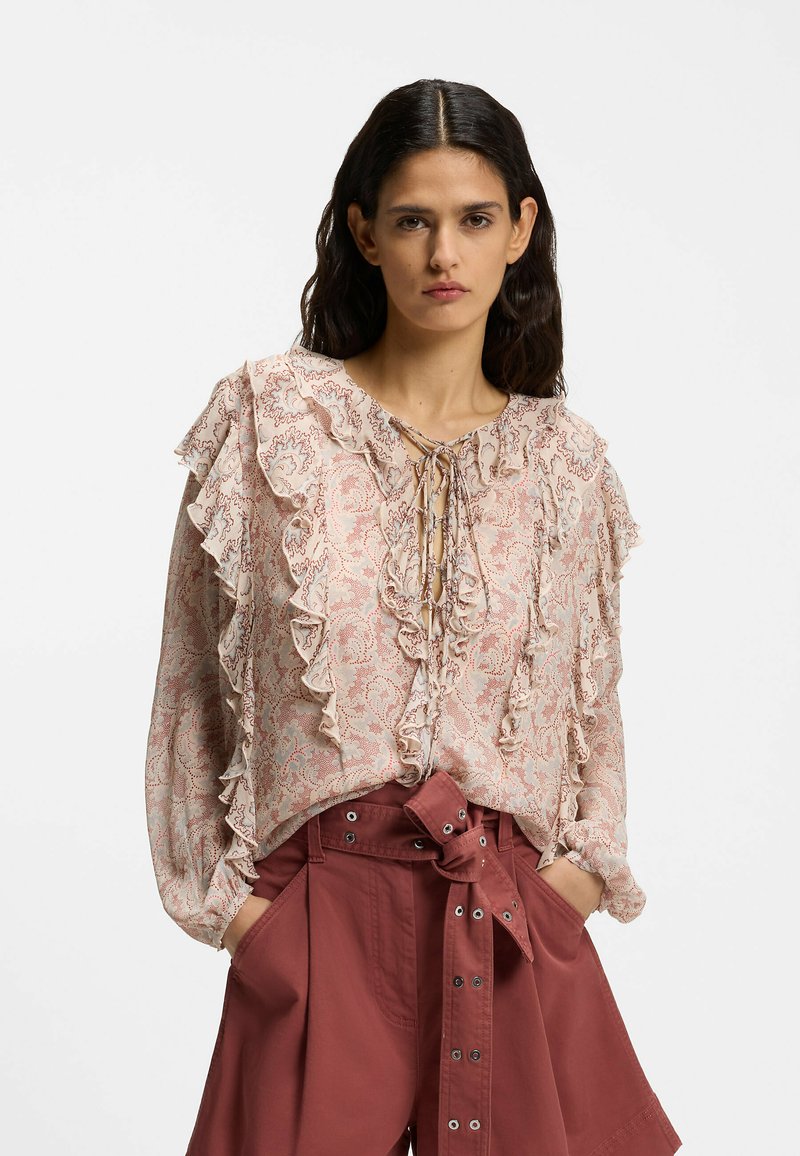 Femme portant une blouse à volants à motifs avec un col noué et un short taille haute couleur rouille avec une large ceinture, les mains dans les poches.