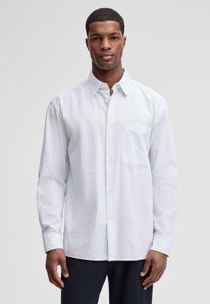 Junger Mann trägt ein hellblau-weiß gestreiftes Hemd mit Brusttasche, steht vor einem schlichten hellgrauen Hintergrund.